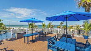 A terrace with blue tables and chairs, umbrellas, and a view of the beach and ocean. - Escape to Paradise–The Beach Club AMI #1 by RVA (Bradenton Beach)