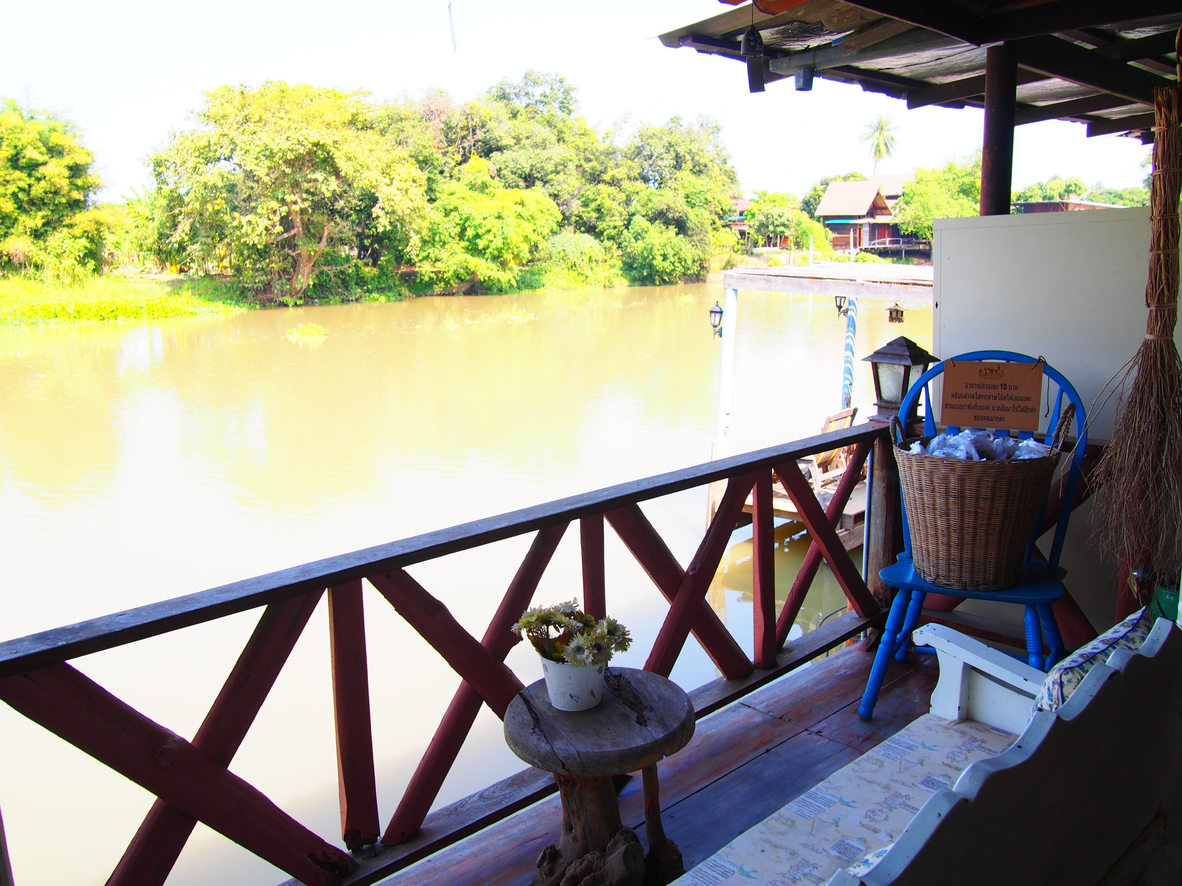 A wooden deck with a railing overlooking a river, a wicker basket, a blue chair, and a small table with a potted plant.