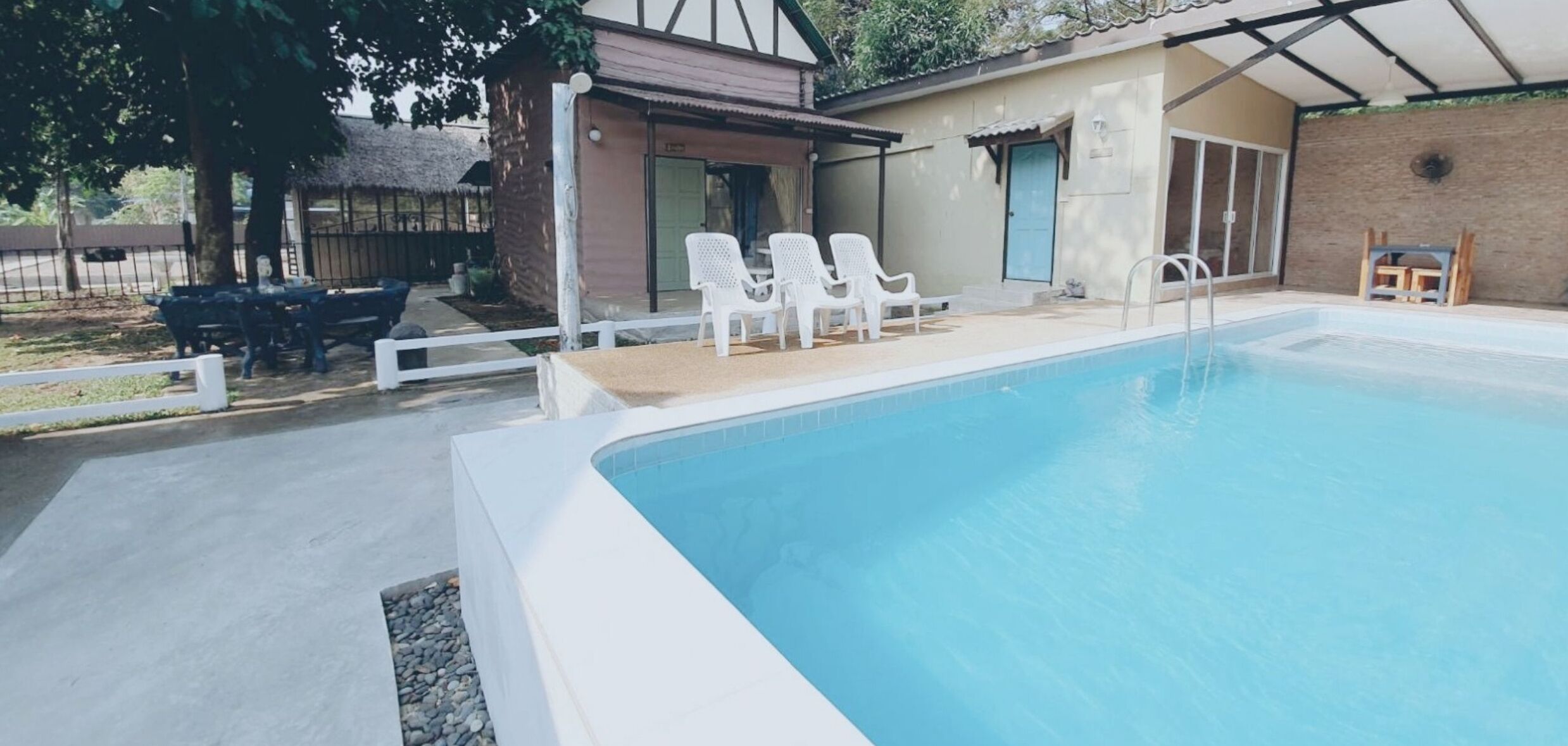 A swimming pool with white lounge chairs, a covered patio area, and a small building with a door and windows.