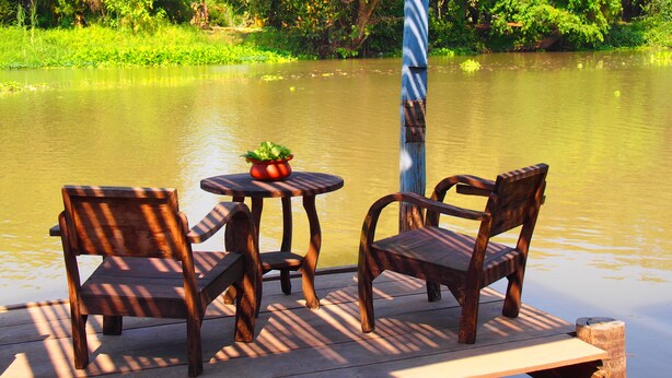 Een houten terras met twee stoelen en een kleine tafel, uitkijkend op kalm water met weelderige begroeiing op de achtergrond.