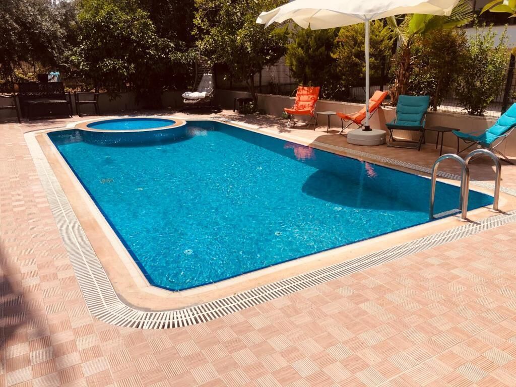 A rectangular swimming pool with a tiled deck, surrounded by lounge chairs and a sun umbrella.