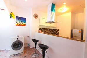 A compact kitchen with a washing machine, two bar stools, a wall-mounted sink, and a clock.