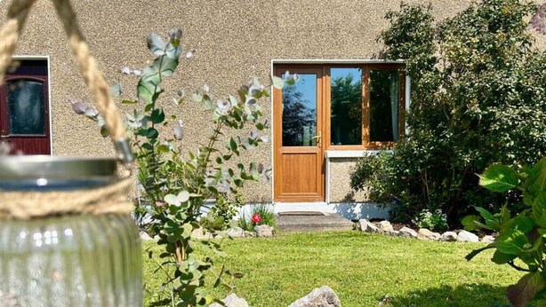 A house with a wooden door, a window, and a garden with rocks and plants. - Charming, Modern Masonette in Fermoy River Town. Pet Friendly. (Fermoy)