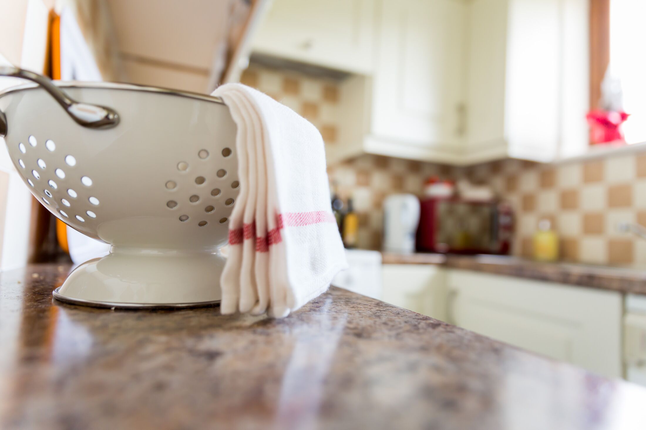 A kitchen with a colander on the counter, a towel, and various kitchen items in the background.