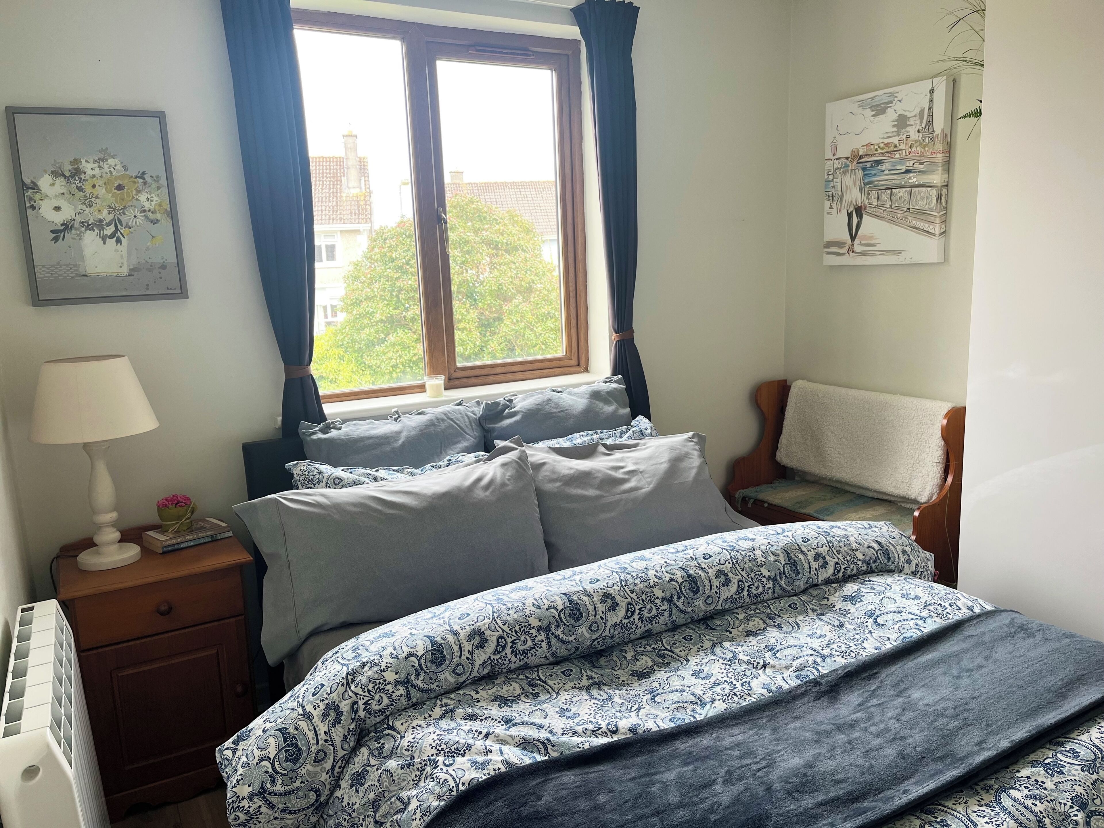 Een slaapkamer met een bed, nachtkastje, lamp, raam met gordijnen en een schilderij aan de muur.