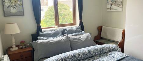 Een slaapkamer met een bed, nachtkastje, lamp, raam met gordijnen en een schilderij aan de muur.