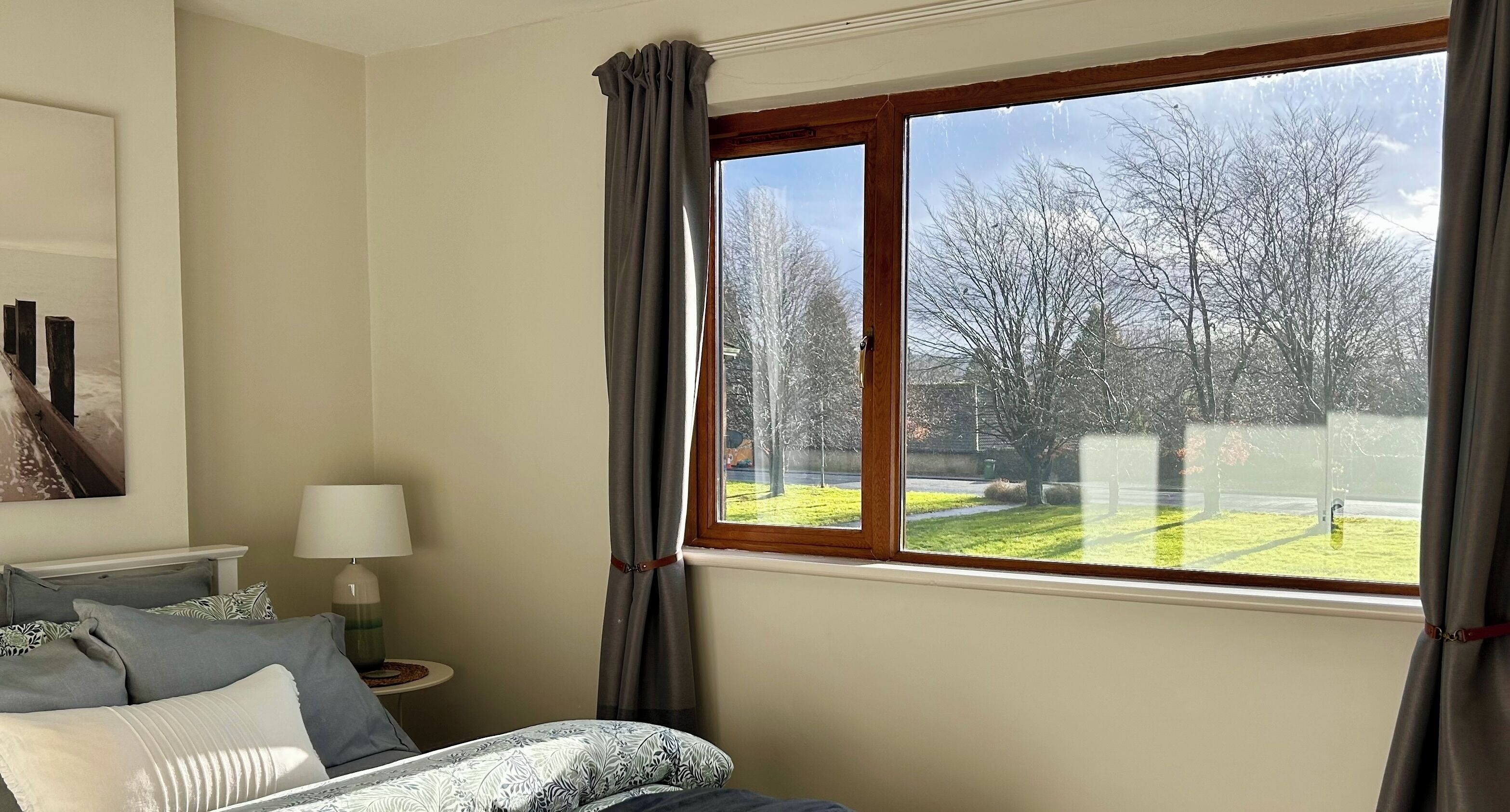 A bedroom with a large window, a bed with patterned bedding, a bedside table with a lamp, and a view of a green landscape with trees.