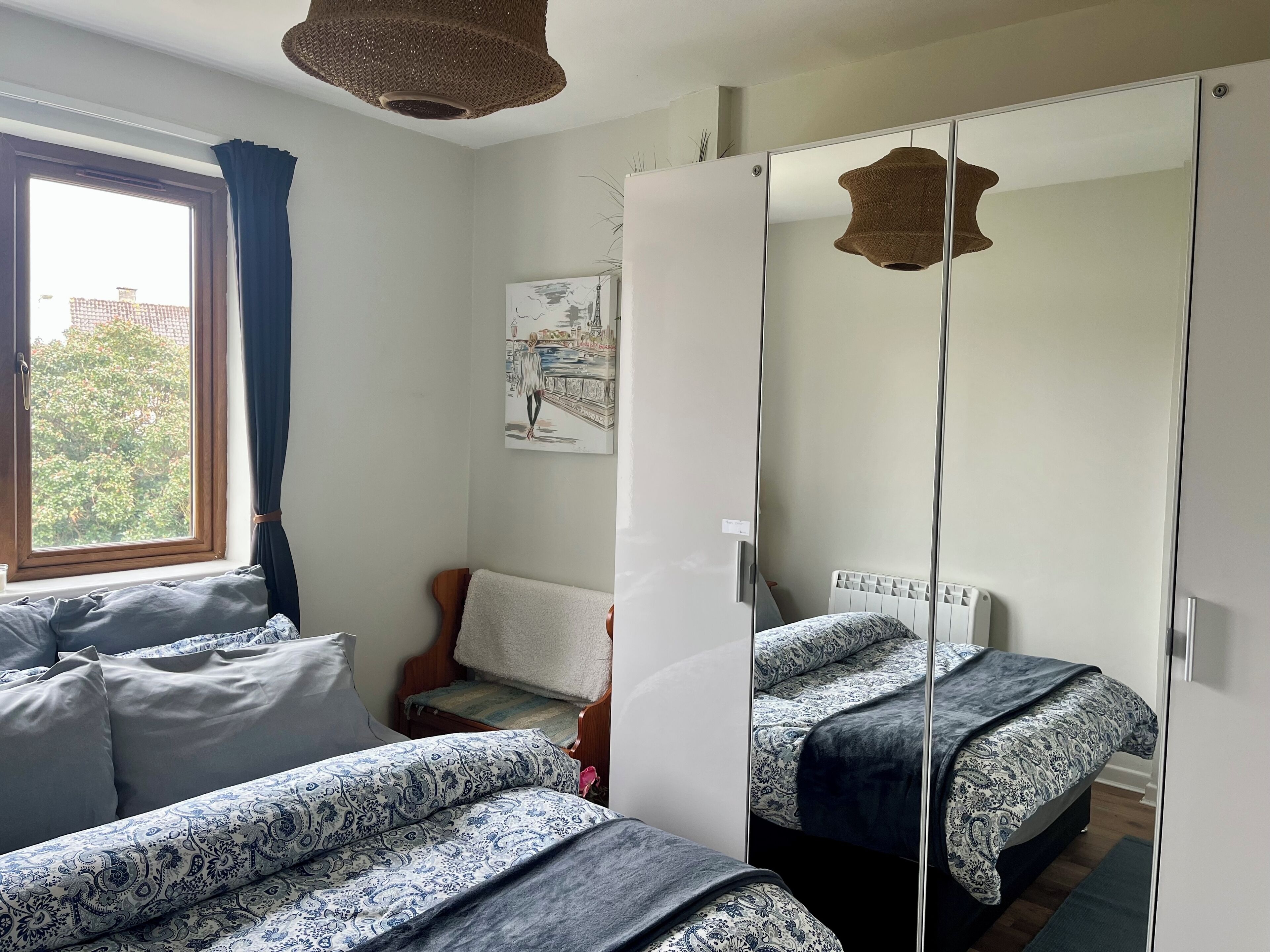 A bedroom with two beds, a window with curtains, a chair, a mirror, and a wall-mounted artwork.