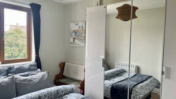 A bedroom with two beds, a window with curtains, a chair, a mirror, and a wall-mounted artwork.