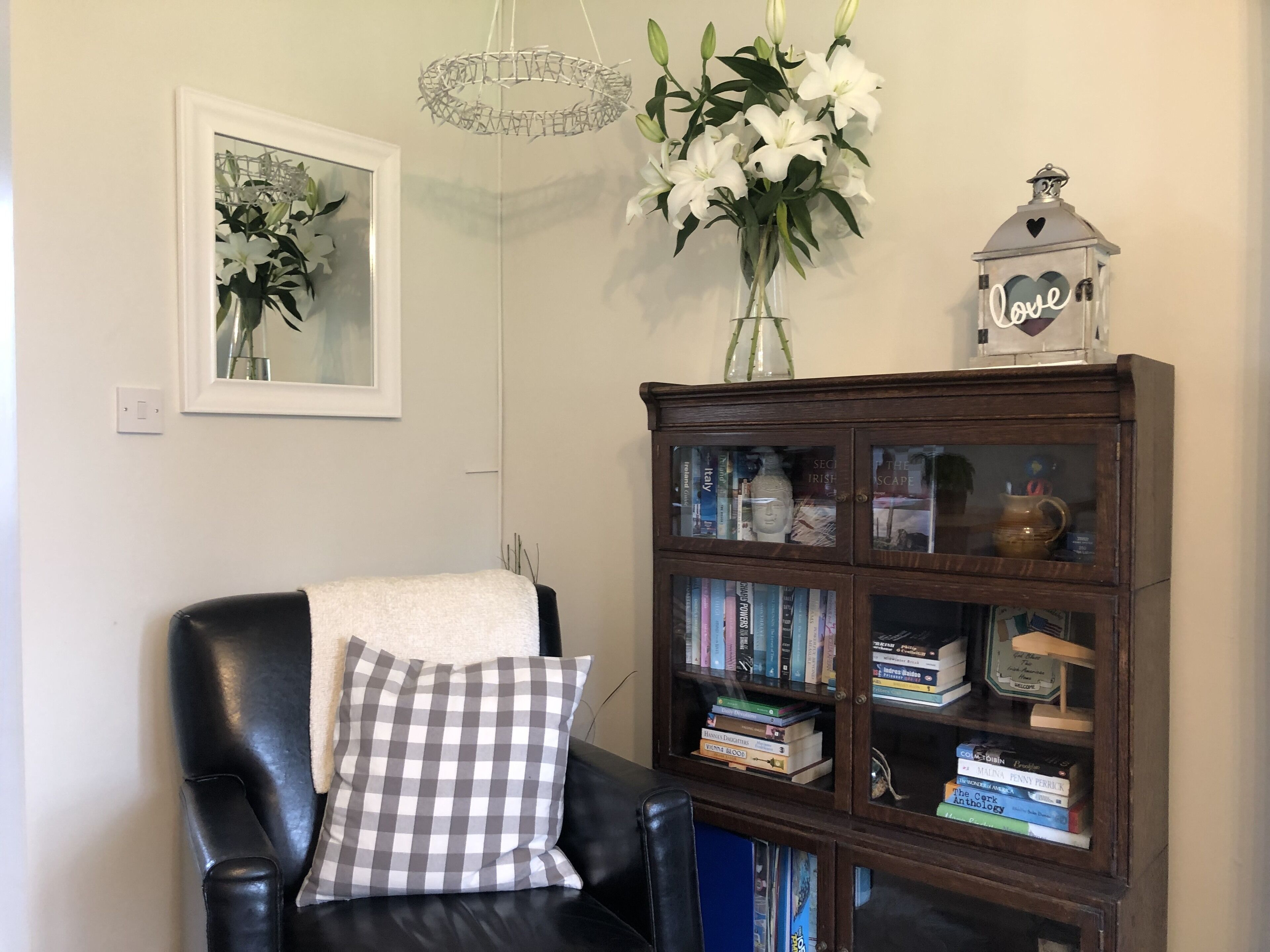 A living room with a leather chair, a checkered pillow, a bookshelf, a framed picture, and a vase with white flowers.
