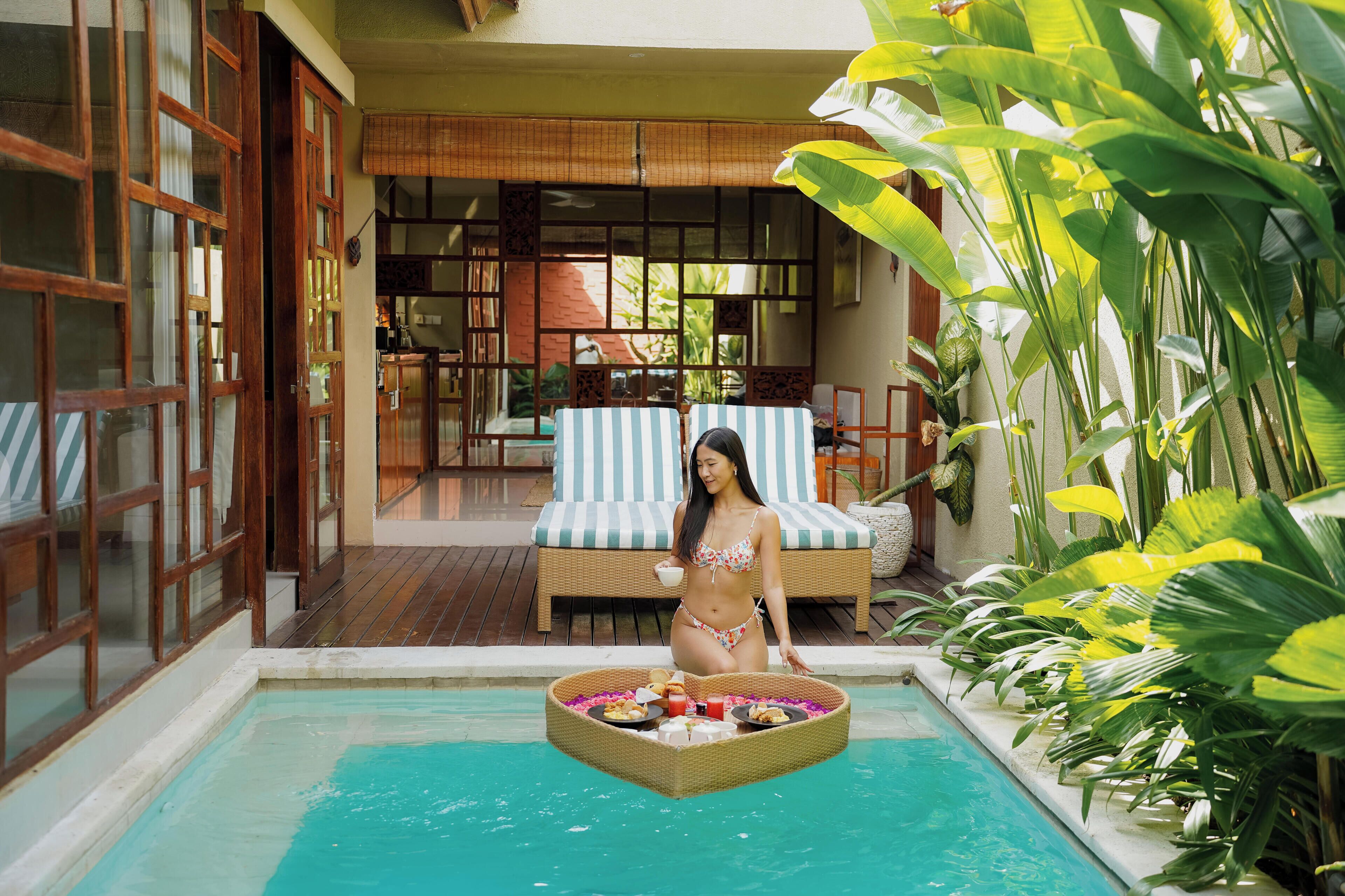 A woman sitting by a pool with a floating tray of food, surrounded by lush greenery and a traditional-style room with a view of the outdoors.