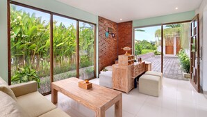 A spacious living area with a large sliding glass door leading to a garden, a wooden coffee table, a sofa, and a wooden cabinet with shelves. - Manca Villa Canggu by Ini Vie Hospitality (Canggu)