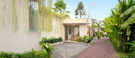 A narrow pathway leading to a building with a glass door, surrounded by lush greenery and a clear blue sky.