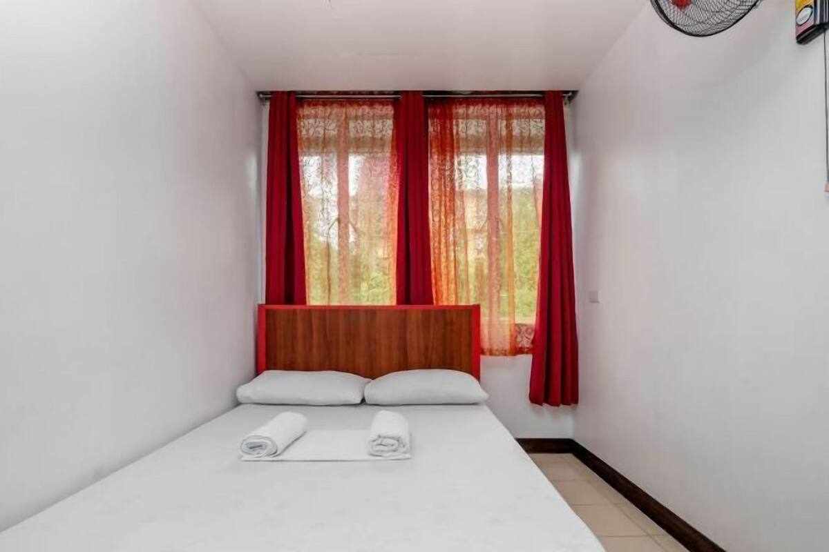 A small, single-bed room with a wooden headboard, red curtains, and white towels on the bed.