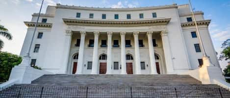 A large, white building with a grand entrance, steps leading up to it, and a fence in the foreground.
