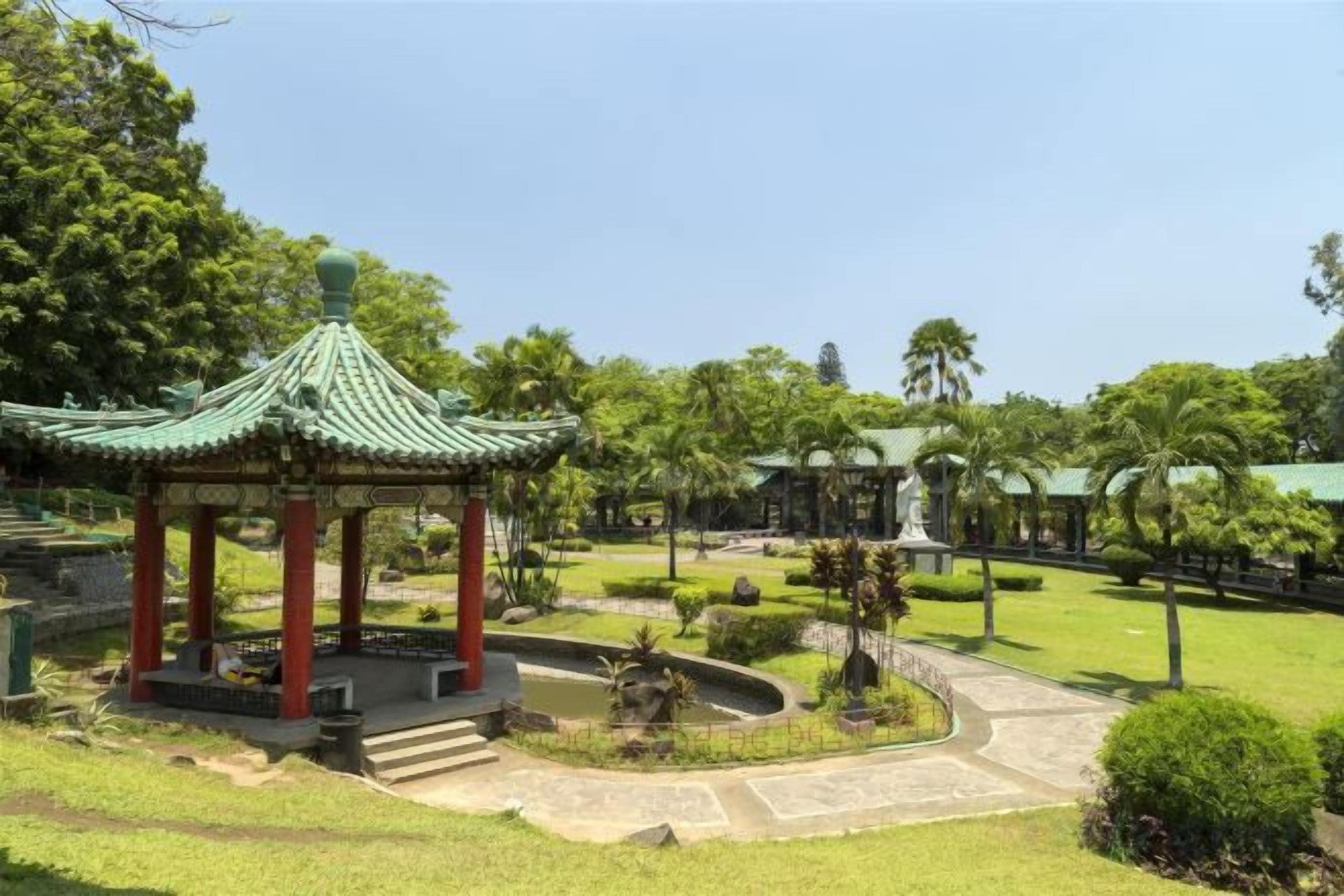 A traditional pavilion with a green roof, surrounded by a well-maintained garden with a curved pathway and a small pond.
