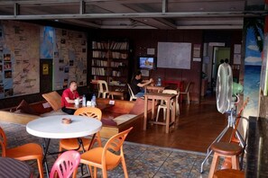Ein gemütlicher Innenbereich mit Tischen, Stühlen, einem Bücherregal und einem Fernseher.