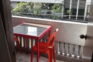 A balcony with a glass table and two chairs, a metal railing, and a view of buildings and greenery.