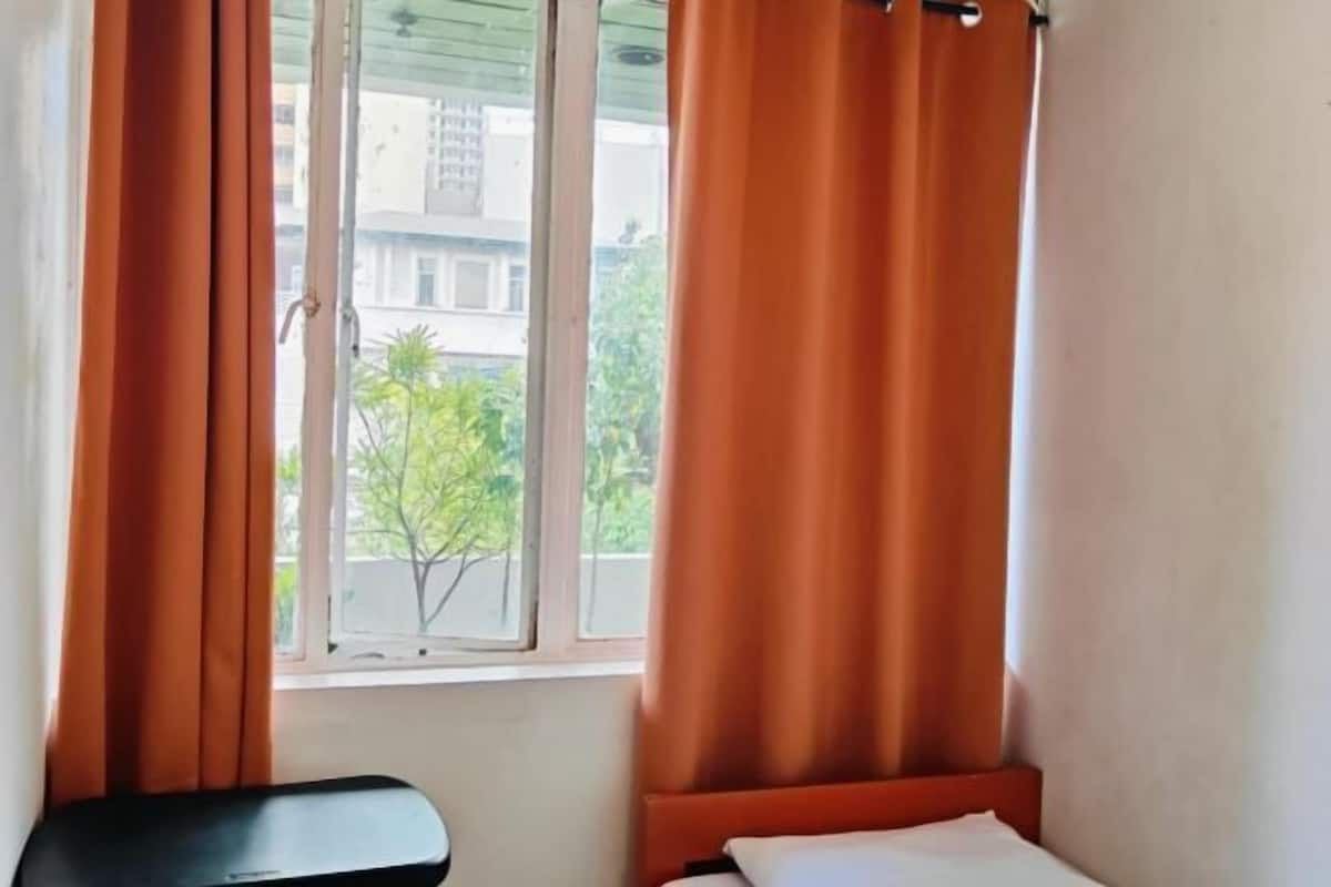A room with a bed, a window with curtains, and a view of buildings and trees outside.