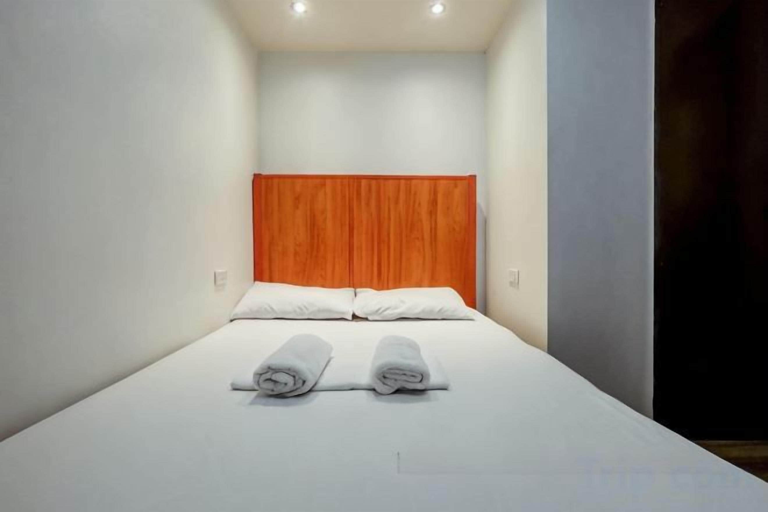 A small, single-bed room with a plain white wall and a wooden headboard.