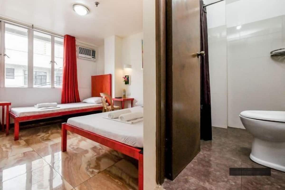 A room with a bed, a small table, a window with a red curtain, and a bathroom with a toilet and shower.