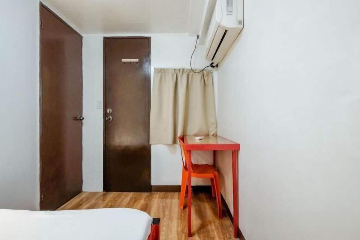 A small room with a bed, a desk, a chair, a door, and an air conditioning unit.