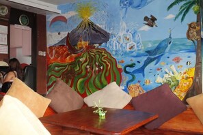 Una habitación con un mural que representa un volcán y una escena oceánica, una mesa de madera con cojines y vistas a la calle.