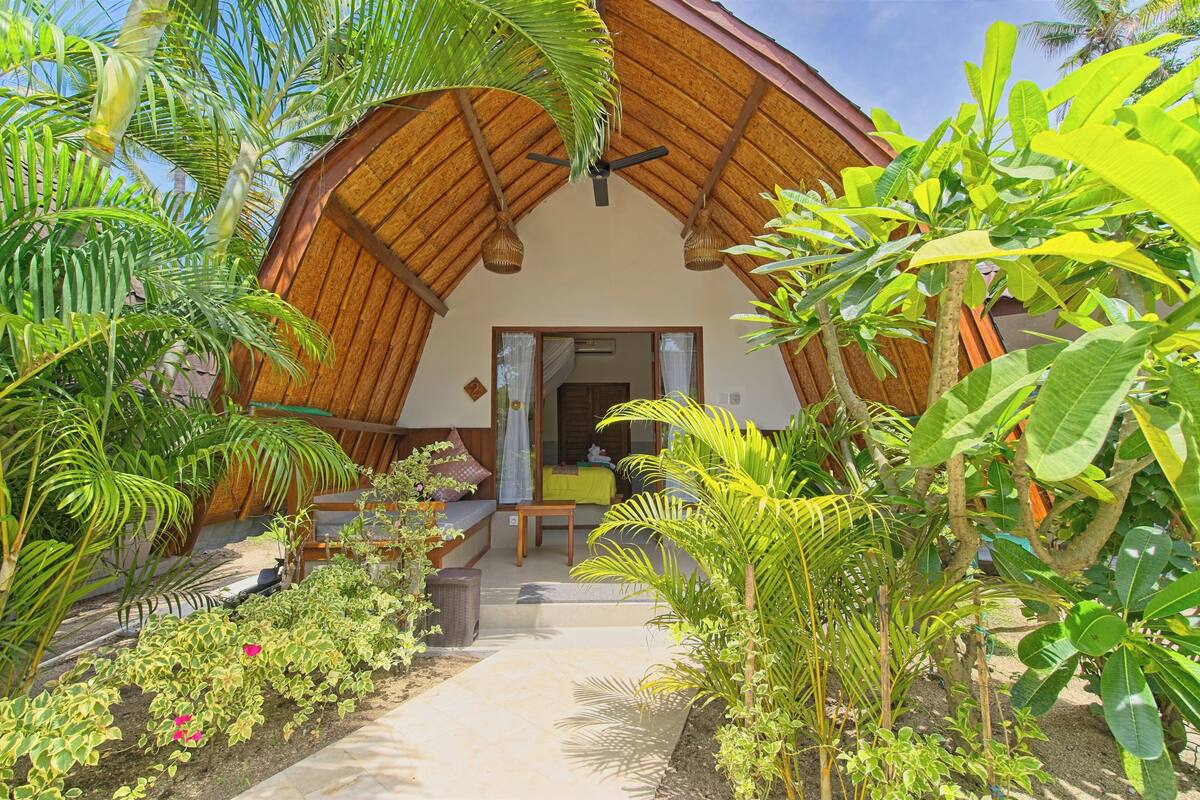 A thatched-roof bungalow with a covered porch, surrounded by lush tropical plants.