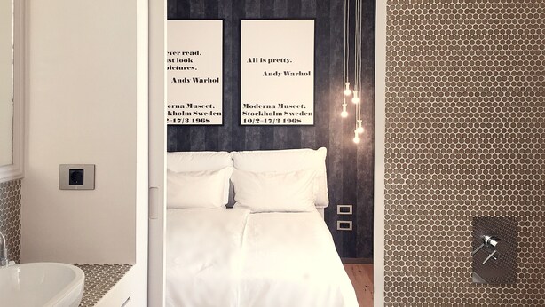 Une chambre moderne avec un grand lit, un mur blanc et un mur à motifs. On y trouve une fenêtre avec des rideaux, un luminaire fixé au mur et une petite étagère.