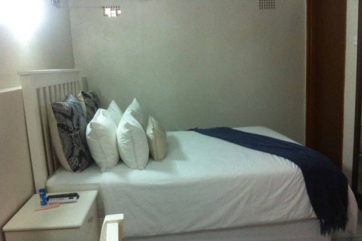 A single bed with white bedding and a blue throw blanket, a bedside table with a lamp, and a patterned pillow.