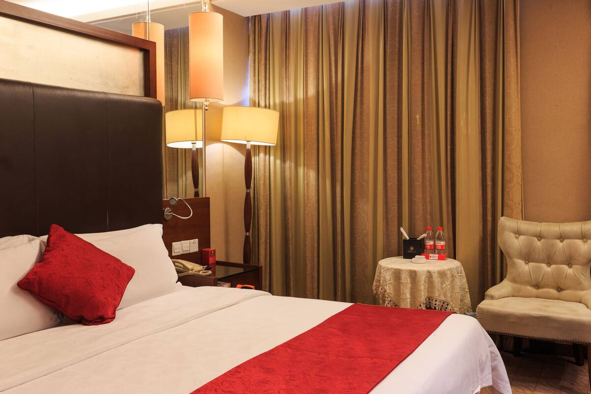 Une chambre d’hôtel avec un grand lit, un plaid rouge, une chaise, une petite table avec une lampe et des rideaux.