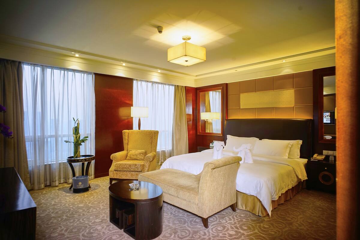 Une chambre d’hôtel avec un grand lit, deux fauteuils, une petite table et une plante en pot.