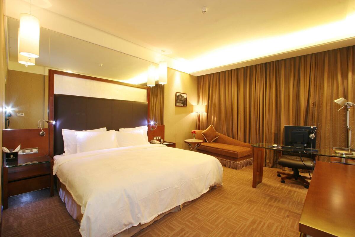 Une chambre d’hôtel avec un grand lit, un bureau avec un ordinateur, une chaise, une petite table avec une lampe et une grande fenêtre avec des rideaux.