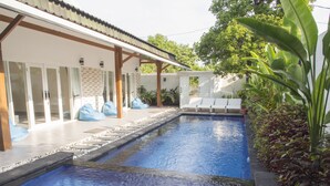 A swimming pool with two lanes, surrounded by lounge chairs and bean bags, with a covered patio area and plants. - KyGunAya Villa (Gili Air)