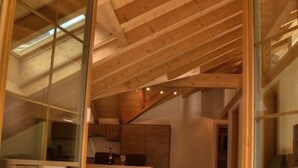 Een ruime loft met hoge plafonds, zichtbare houten balken en grote glazen deuren die leiden naar een eethoek met een rustiek koperen vloerkleed.