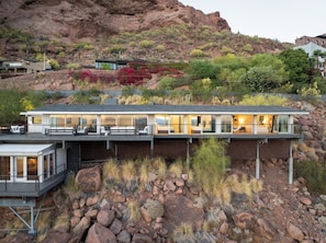 A modern house with a flat roof, large windows, and a balcony overlooking a rocky landscape.