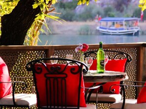 A table with a bottle and glasses, red chairs, and a view of a boat on the water. - Aydos Club (Ortaca)