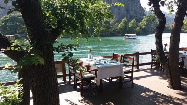 A wooden deck with tables and chairs overlooking a body of water, with a boat visible in the distance. - Aydos Club (Ortaca)