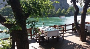 Une terrasse en bois, agrémentée de tables et de chaises, donnant sur une étendue d’eau, avec un bateau visible au loin.