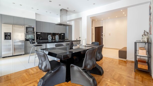 A modern kitchen with stainless steel appliances, a dining area with a glass table and black chairs, and a wooden floor.
