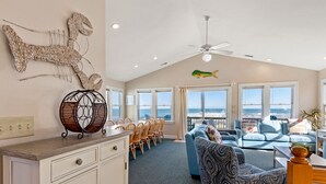A spacious living room with a high ceiling, a ceiling fan, and a large window overlooking the ocean. There is a dining area with wooden chairs and a table, and a blue sofa with patterned cushions. - Ocean's Edge: Oceanfront, Pool, Spa, Dogs OK (Nags Head)