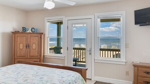 A bedroom with a bed, a ceiling fan, a wooden dresser, and two windows with a view of the beach. - Ocean's Edge: Oceanfront, Pool, Spa, Dogs OK (Nags Head)
