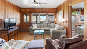 Interior de una cabaña de madera con televisor, sofá y vista a la playa a través de amplias ventanas.