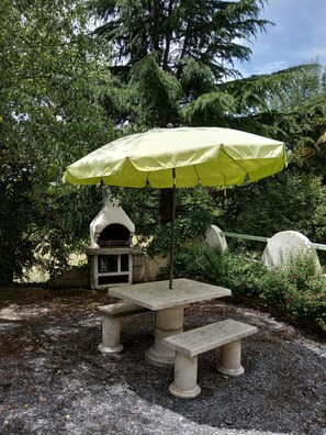Ein Garten mit einem Steintisch und einer Bank unter einem gelben Sonnenschirm, umgeben von Bäumen und einer kleinen Steinstruktur.