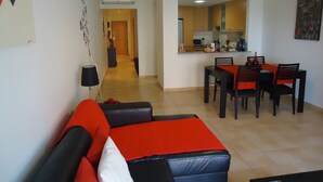 Una sala de estar moderna con sofá, mesa de comedor y sillas. Se ve un área de cocina a través de un vano abierto.