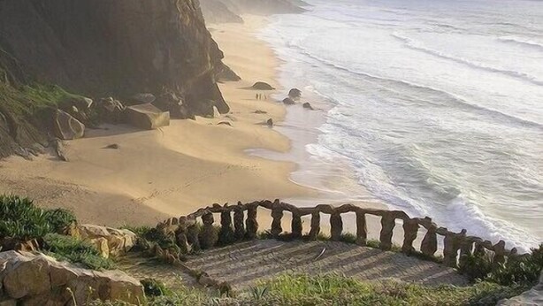 Un paisaje costero con una playa arenosa, acantilados rocosos y una escalera de piedra que baja hacia la orilla.