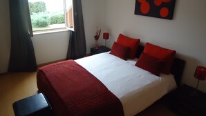 Un dormitorio con una cama grande, una ventana con cortinas, una obra de arte en la pared y una mesita de noche con una vela roja.