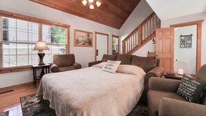 A bedroom with a large bed, a ceiling fan, wooden beams, a staircase, and a window with blinds.