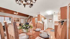 A dining area with a wooden table set for four, surrounded by wooden chairs. A chandelier hangs from the ceiling, and there is a kitchen area visible in the background.