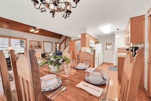 Een eetgedeelte met een houten tafel voor vier personen, omringd door houten stoelen. Een kroonluchter hangt aan het plafond, en er is een keuken te zien op de achtergrond.